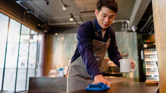 Barista wearing an apron cleaning counter while holding a coffee mug in a modern cafe setting.