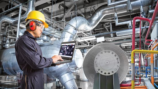 Man in hardhat typing on laptop in an interior industrial setting.