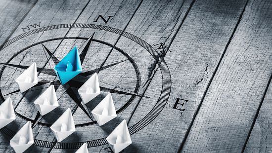 Blue Paper Boat Leading A Fleet Of Small White Boats With Compass Icon On Wooden Table With Sunlight